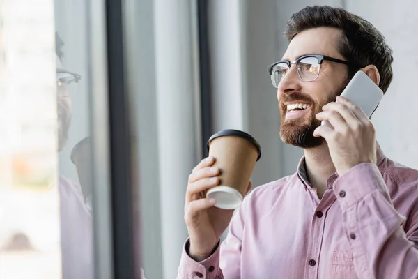 Happy manager in shirt talking on mobile phone and holding coffee to go in office — Stock Photo