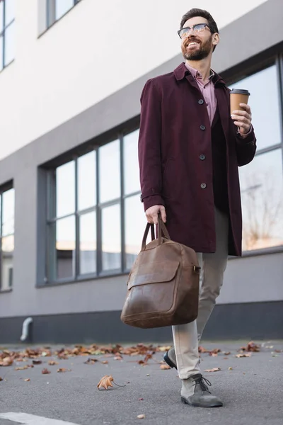 Веселий бізнесмен в траншеї тримає сумку і каву, щоб піти на міську вулицю — стокове фото