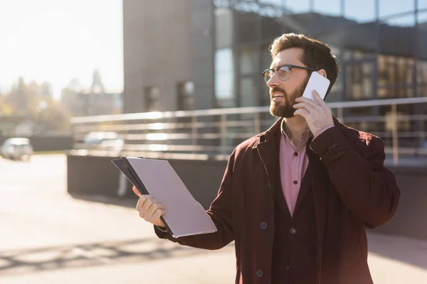 Uomo d'affari positivo che parla su smartphone e tiene cartelle di carta sulla strada urbana — Foto stock