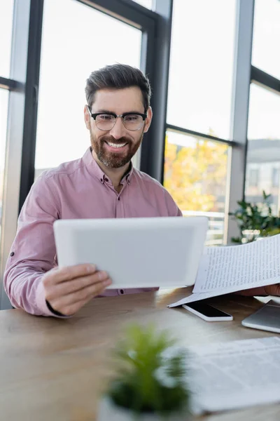 Uomo d'affari felice guardando tablet digitale vicino a carte e gadget in ufficio — Foto stock