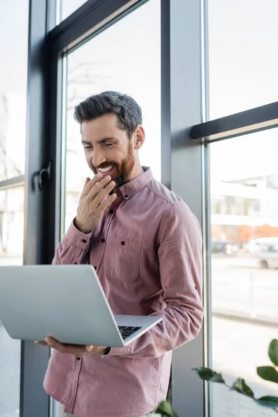 Homme d'affaires heureux en chemise tenant ordinateur portable près de la fenêtre dans le bureau — Photo de stock