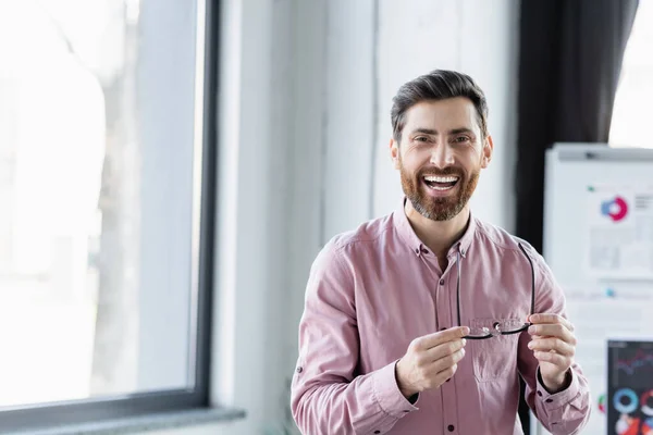 Happy businessman in shirt holding eyeglasses and looking at camera in office — Stock Photo