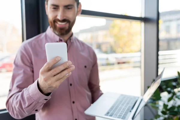 Smartphone en la mano del empresario borroso con el ordenador portátil en la oficina - foto de stock