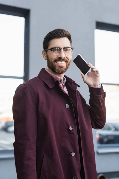 Positiver Geschäftsmann im Trenchcoat blickt in die Kamera, während er sein Handy mit leerem Bildschirm im Freien hält — Stockfoto