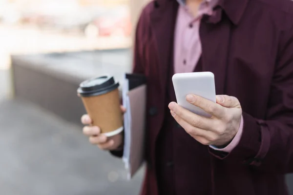Cropped view of blurred businessman with paper cup and folders holding smartphone outdoors — Stock Photo