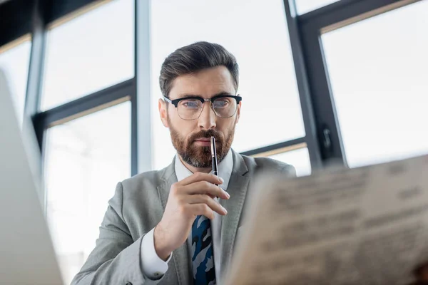 Бородатый бизнесмен держит ручку и читает газету возле компьютера в офисе — стоковое фото