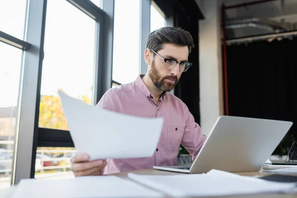 Empresario en anteojos trabajando con papeles y laptop en oficina - foto de stock