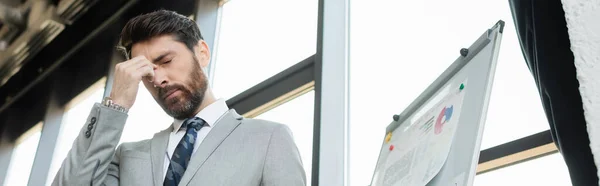 Low angle view of tired businessman in suit standing near flip chart in office, banner — Stock Photo