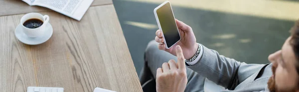 Vue grand angle d'un homme d'affaires flou pointant vers un smartphone près d'un journal et d'un café au bureau, bannière — Photo de stock