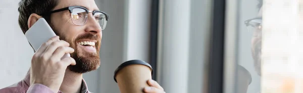 Positive businessman in eyeglasses talking on smartphone and holding takeaway drink in office, banner — Stock Photo