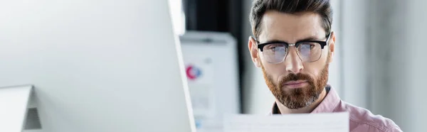 Manager in eyeglasses looking at paper near computer monitor in office, banner — Stock Photo