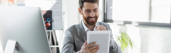 Улыбающийся бизнесмен с цифровым планшетом возле монитора компьютера в офисе, баннер — стоковое фото