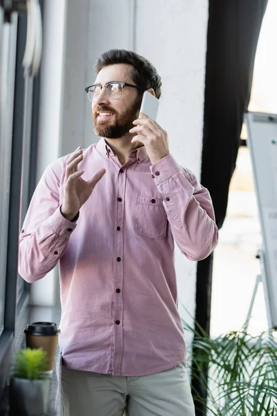 Homme d'affaires en chemise parlant sur smartphone près du café pour aller sur le rebord de la fenêtre dans le bureau — Photo de stock