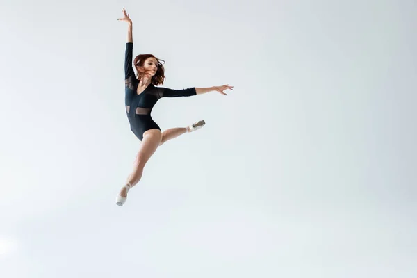 Longitud completa de bailarina en traje negro saltando aislado en gris - foto de stock