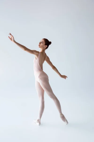 Повна довжина балерини в боді-костюмі танцює з витягнутою рукою на сірому — стокове фото
