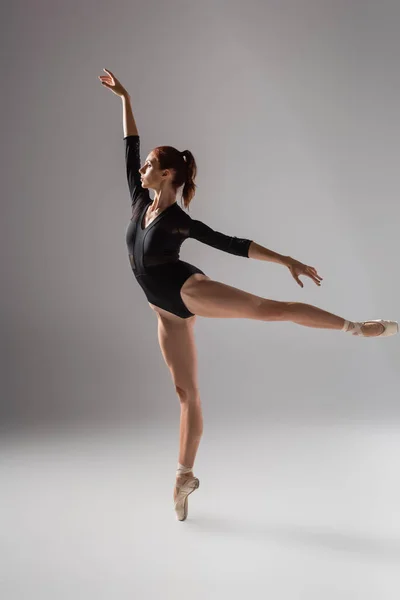 Full length of elegant ballerina in black bodysuit dancing with raised hand on dark grey — Stock Photo