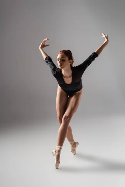 Full length of pretty ballerina in black bodysuit dancing with raised hands on dark grey — Stock Photo