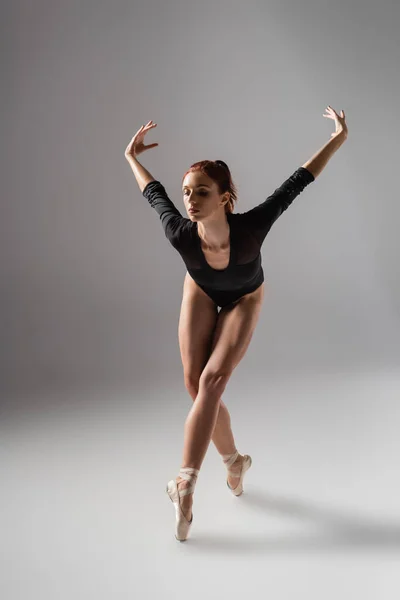 Larga duración de bonita bailarina en traje negro bailando con las manos levantadas sobre gris oscuro - foto de stock