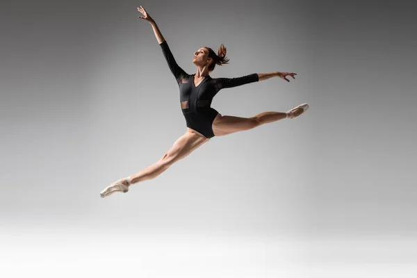 Полная длина молодой балерины в пуантах и черных телесных костюмах, прыгающих на темно-сером — стоковое фото