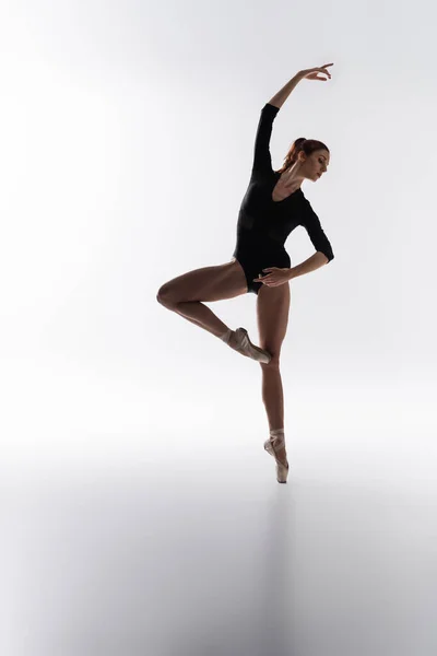 Larga duración de la joven bailarina en zapatos puntiagudos y body negro bailando sobre gris - foto de stock