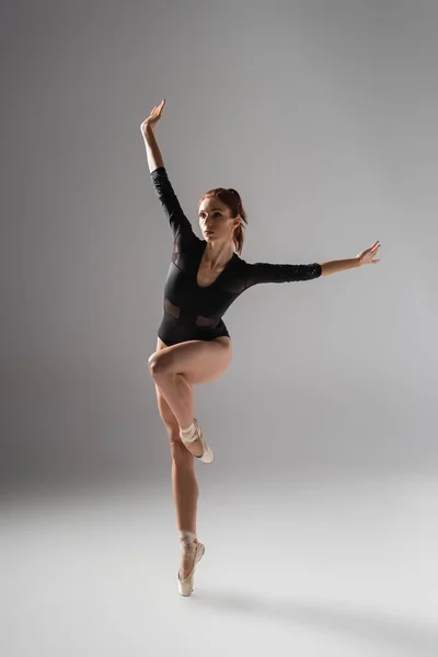 Повна довжина витонченої балерини в чорному комбінезоні танцює з витягнутими руками на темно-сірому — Stock Photo