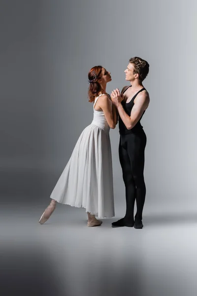 Full length of graceful woman in white dress holding hands in ballet dancer on dark grey — Stock Photo