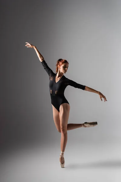 Graceful ballerina in pointe shoes and black bodysuit with outstretched hands on dark grey — Stock Photo