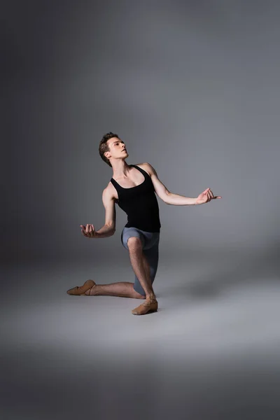 Полная длина элегантной балерины, стоящей на коленях, исполняя балетный танец на темно-сером — стоковое фото