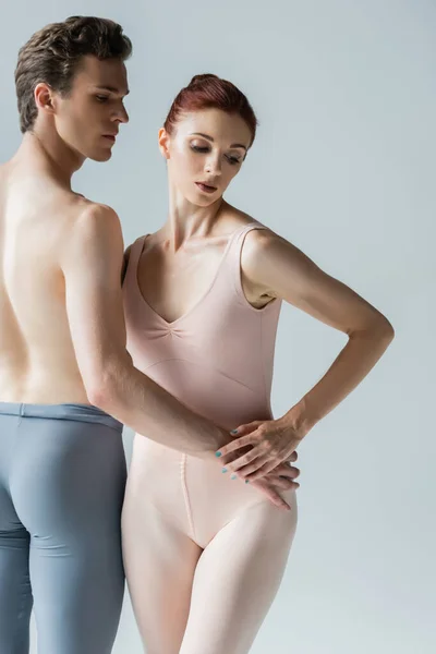 Танцовщица без рубашки держит за руку бедро молодой балерины, изолированной на сером — стоковое фото