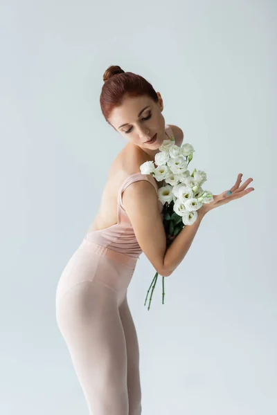 Redhead ballerina with closed eyes holding bouquet of eustoma flowers isolated on grey — Stock Photo