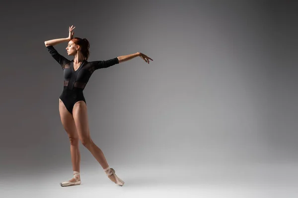 Повна довжина витонченої балерини в чорному комбінезоні, що стоїть з витягнутою рукою на темно-сірому — стокове фото