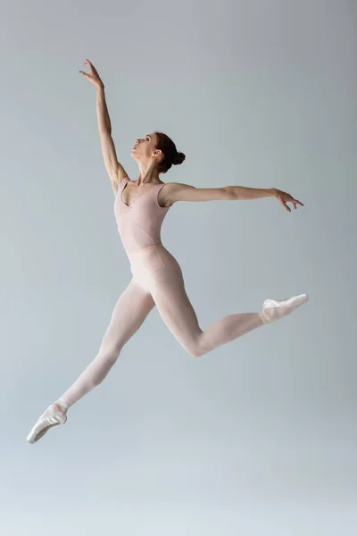 Longitud completa de elegante bailarina en traje de cuerpo levitando y haciendo gestos en gris - foto de stock