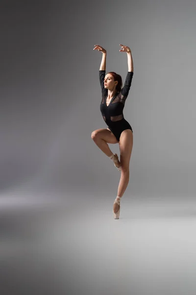 Bailarina joven y elegante en zapatos puntiagudos y body negro con las manos extendidas sobre gris oscuro - foto de stock