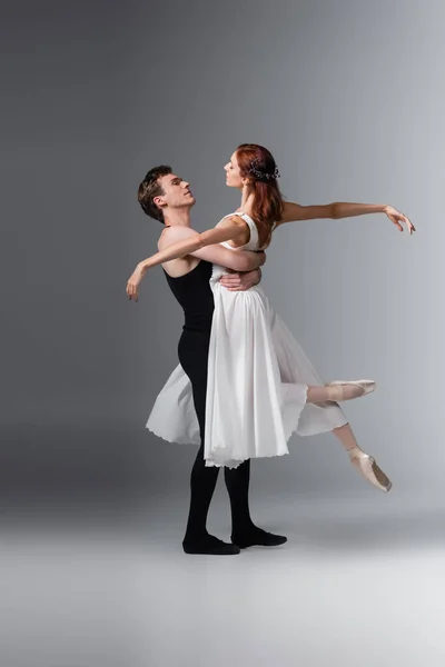 Comprimento total do homem levantando bailarina graciosa em vestido branco no cinza escuro — Fotografia de Stock