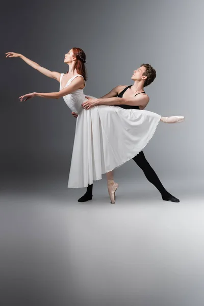 Comprimento total de bailarina graciosa no vestido branco dançando com o jovem parceiro no cinza escuro — Fotografia de Stock