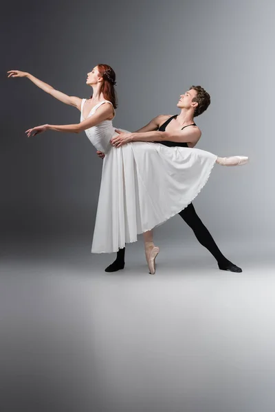 Full length of graceful ballerina in white dress dancing with young partner on dark grey — Stock Photo