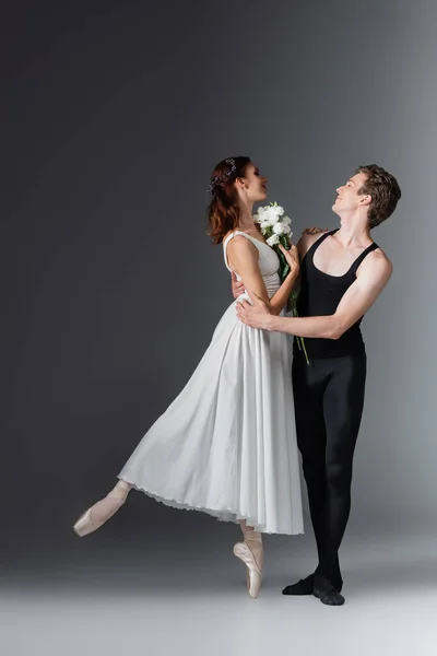 Full length of graceful ballerina in white dress holding flowers and dancing with partner on dark grey — Stock Photo