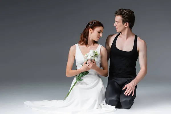 Graceful ballerina in white dress holding flowers and standing on knees with partner on dark grey — Stock Photo