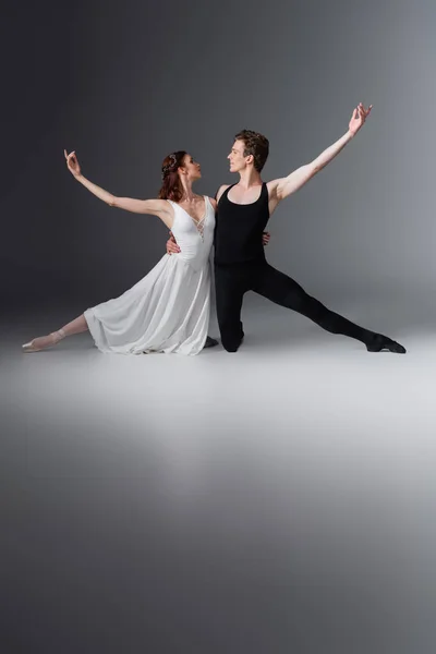 Pleine longueur de gracieuse ballerine en robe blanche dansant avec partenaire se regardant sur le gris foncé — Photo de stock