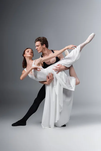 Longitud completa del hombre apoyando elegante bailarina en vestido blanco mientras baila en gris oscuro - foto de stock