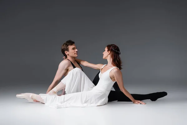 Повна довжина витонченої балерини в білій сукні, сидячи з усміхненим партнером на темно-сірому — стокове фото