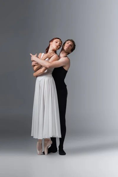 Longueur totale de danseuse de ballet embrassant gracieuse ballerine en robe blanche sur gris foncé — Photo de stock