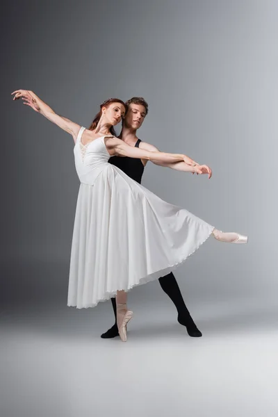 Pleine longueur de gracieuse ballerine en robe blanche dansant avec jeune partenaire sur gris foncé — Photo de stock