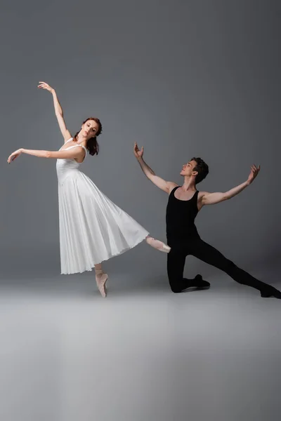 Longitud completa de elegante bailarina en vestido blanco bailando cerca de la pareja de pie sobre la rodilla en gris oscuro - foto de stock