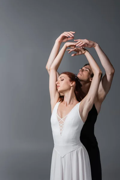 Graceful ballerina in white dress dancing with partner while raising hands on dark grey — Stock Photo