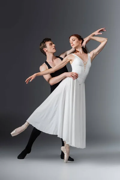 Pleine longueur de jeune ballerine gracieuse en robe blanche dansant avec son partenaire sur gris foncé — Photo de stock