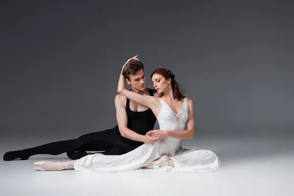 Full length of flexible dancer and graceful ballerina in white dress sitting on dark grey — Stock Photo