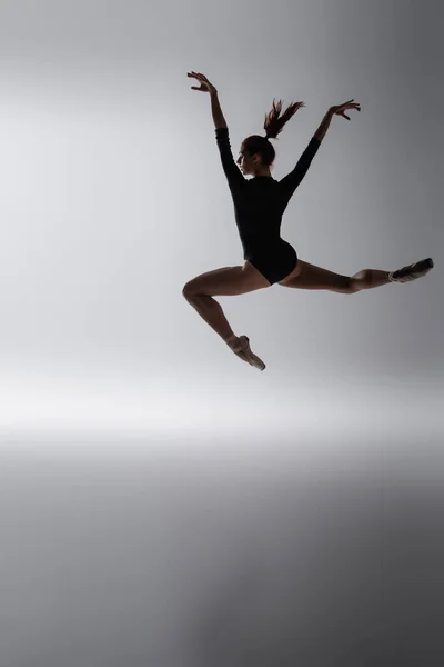 Redhead ballerina in bodysuit jumping with outstretched hands on dark gray — Stock Photo