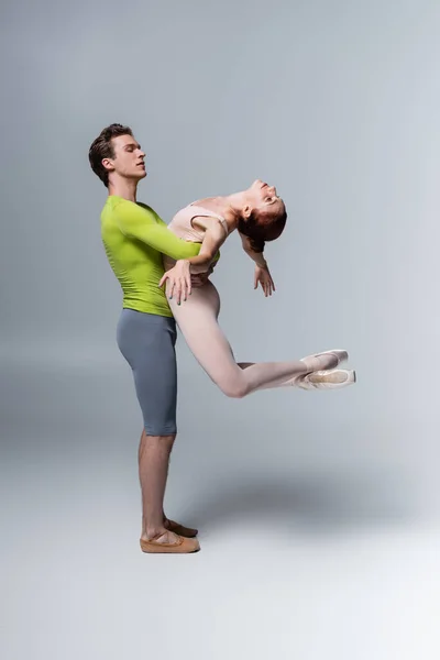 Bailarina de ballet fuerte levantando bailarina en traje de cuerpo en gris - foto de stock
