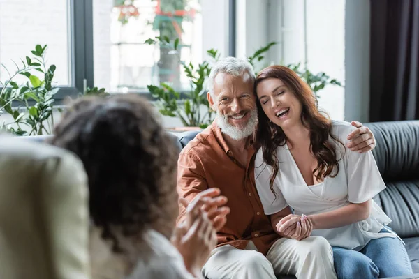 Fröhlicher Mann umarmt glückliche Frau während Beratung mit verschwommenem afrikanisch-amerikanischem Psychologen — Stockfoto